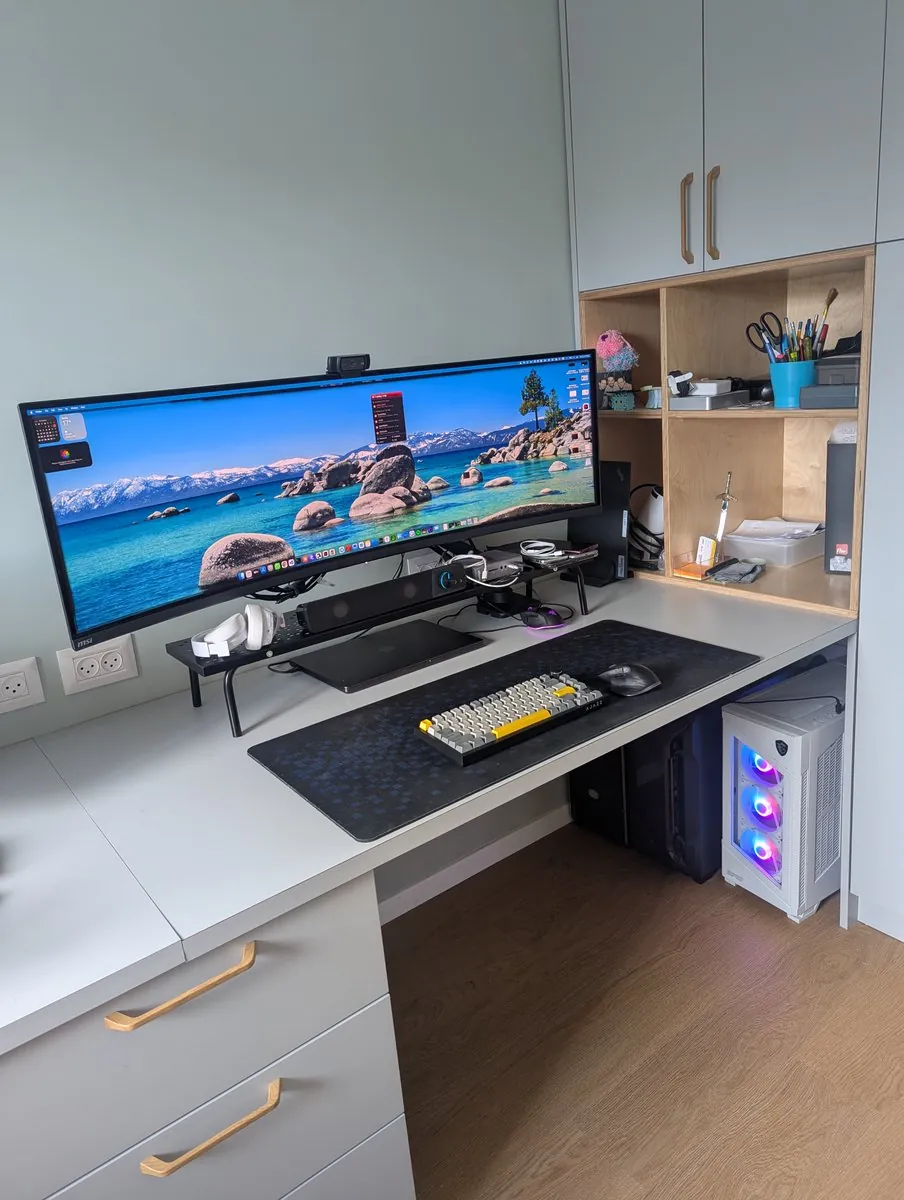 The full corner: the ultrawide floating on its arm over a clear desk, shelf unit beside it, and a white desktop tower with RGB fans below